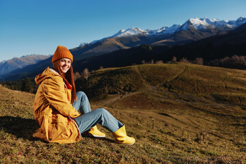 Woman sitting on a hill on the grass rest smile with teeth looking at the mountains in the snow in the winter in a yellow raincoat and jeans happy sunset trip on a hike, freedom lifestyle 
