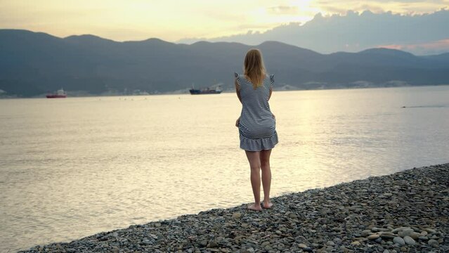 slender pretty young woman is preparing to swim in sea or ocean in bay and takes off her striped dress. girl in lilac swimsuit is ready to swim in natural pond during her journey.