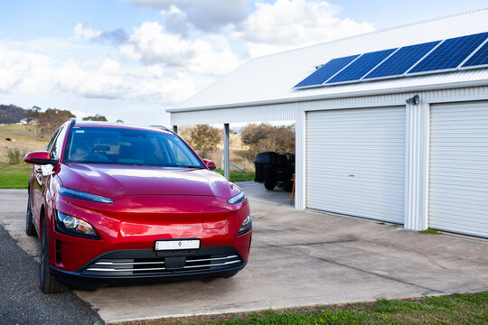 Red electric car beside shed garage with solar panels on roof in country