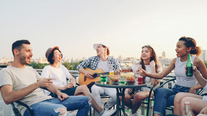 Creative guy in casual clothes is playing the guitar and his friends multi-ethnic group are singing holding bottles with beer and soft drinks sitting in circle at table on roof.