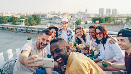 Happy young men and women are taking selfie with smart phone posing, laughing and making hand gestures celebrating holiday sitting on roof at table with food and drinks.