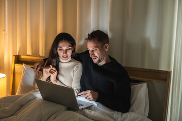 Caucasian couple sitting on their bed under blanket in dark room watching laptop screen.