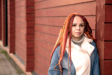 Portrait of young woman with ginger dreadlocks standing leaning on wall, on city street. Positive female looking at camera, smiling.