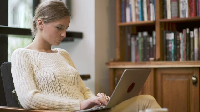 Young beautiful woman is sitting with laptop computer on her knees and typing on the keyboard. Lady is serious and focused on task. She is writing letter, book, project description.