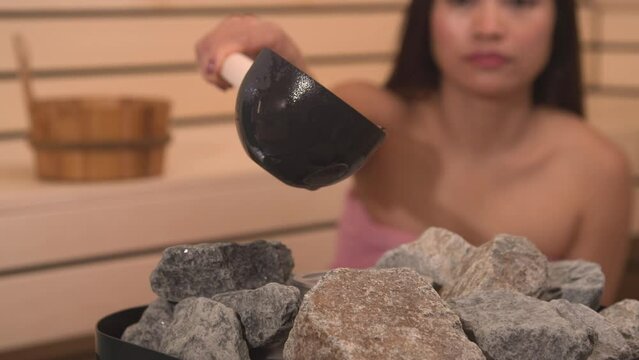 CLOSE UP: Woman pouring water to heater stones to make steam in Finnish sauna. Stones as necessary equipment for slightly slower evaporation of sauna steam. Beautiful lady treating herself with spa.