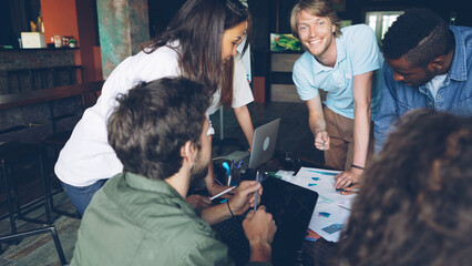 Group of young people colleagues is discussing charts and graphs standing around table in modern office. Business, successful teamwork and professionals concept.