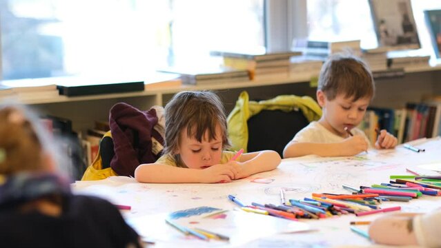 Cute little children draw colorful pictures with markers sitting at long table near bookshelves in contemporary kindergarten