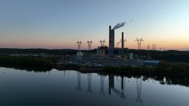 Factory power plant generates electricity. Towers with electric cables reflect in river water at sunset. Rising aerial.