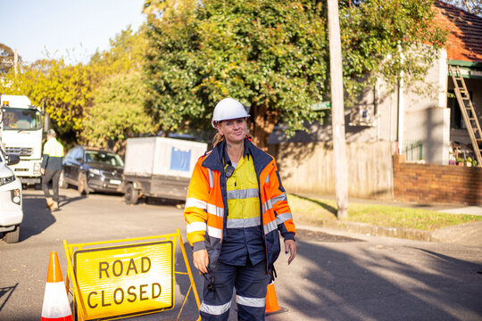 Female road worker wearing white helmet and orange jacket on the road with a road closed sign
