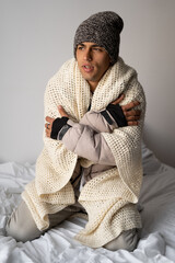 Young man warmly dressed is feeling cold, covered with warm blanket, frozen sitting on the bed at...
