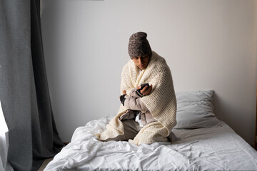 Young man warmly dressed is feeling cold, covered with warm blanket, frozen sitting on the bed at...