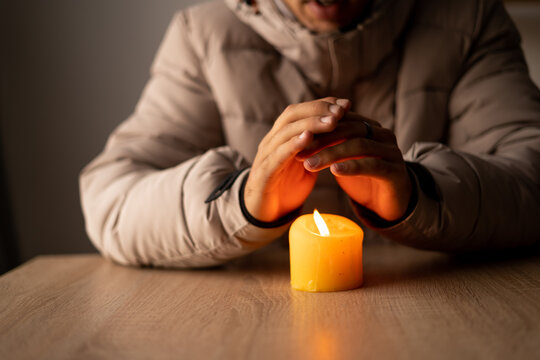 A man warms his hands from a burning candle at cold house, close up. Shutdown of heating and electricity, power outage, blackout, energy crisis.