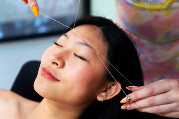 professional beautician performs an eyebrow threading on a young asian woman in the beauty salon,...