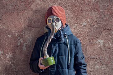 Portrait of a boy in a gas mask and warm clothes against the background of a red old plastered wall. Knitted orange hat and black jacket. Gas mask filter in hand. Dirty texture. Apocalypse concept