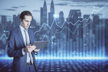 Attractive young european businessman with tablet and abstract glowing blue business graph hologram on blurry city background. Economy, finance and digital money concept. Double exposure.