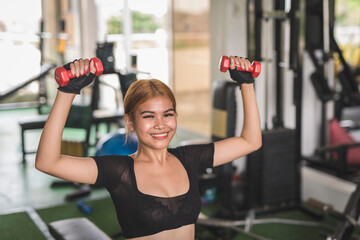 Fototapeta na wymiar A young and sexy asian woman smiles while doing seated dumbbell shoulder presses on a flat bench at the gym. Wearing a black crop top and bike shorts.
