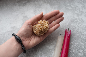 Woman's hand with a black bracelet and a desert rose crystal in the palm on a gray background....