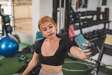 Fototapeta na wymiar A sexy and confident young asian woman poses while taking a selfie of herself during a workout at the gym.