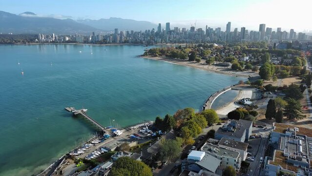 Drone shot of Kitsilano in Vancouver, British Columbia showing residential homes, condo apartment, beach, city skyline, cars on streets, ocean, mountains and trees.