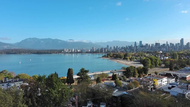 Drone shot of Kitsilano in Vancouver, British Columbia showing residential homes, condo apartment, beach, city skyline, cars on streets, ocean, mountains and trees.