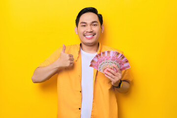 Cheerful young handsome Asian man holding bunch of money and showing thumbs up isolated on yellow background