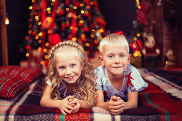 Fototapeta na wymiar Children in pajamas lying on the floor looking at the camera near Christmas tree and fireplace 