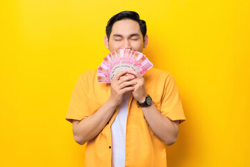 Smiling young handsome Asian man holding bunch of money banknotes and smelling isolated on yellow background