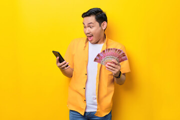 Surprised young handsome Asian man holding bunch of money and using mobile phone isolated on yellow...