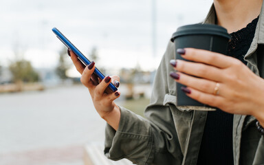 Side view of woman using smartphone during coffee break outdoors. Close-up of female hands holding disposable cup of coffee to go and mobile phone, selective focus on hand with device