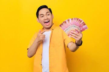Cheerful young handsome Asian man pointing fingers at money banknotes isolated on yellow background