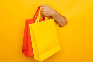 Man holds in hand a yellow and red paper bags for purchases through a hole in yellow background