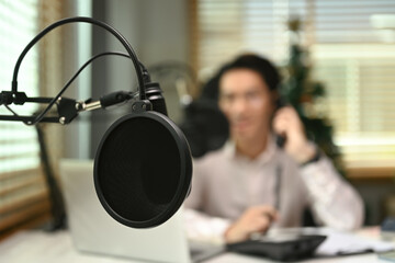 Close up view of a condenser microphone in home studio interior with male radio host sitting in...