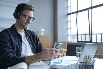 German freelancer guy in headphones sitting at modern home office via laptop while using smartphone