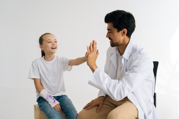 Studio shot of cheerful little injured girl with broken hand wrapped in plaster bandage giving high five to friendly male doctor at checkup meeting, on white isolated background.