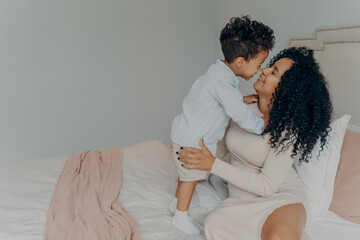 Happy pregnant mother embracing adorable little son while sitting on bed