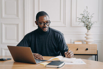 Fototapeta premium Focused young african american businessman writing down notes in notebook while working in office