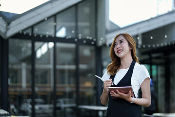 Starting and opening a small business, a young Asian woman showing a smiling face holding a tablet in an apron standing in front of a coffee shop bar counter. Business Owner, Restaurant, Cafe concept