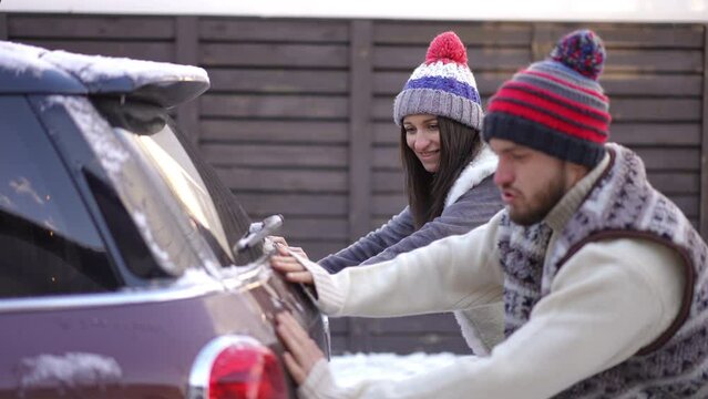 Young Caucasian couple pushing car on winter day outdoors talking. Side view portrait of man and woman travelling on vacations with automobile stuck in snow. Tourism and relationship