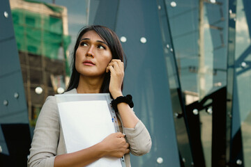 Thai executive woman looking up holding work documents outside office building. Pensive Asian transgender person with files in hand, thinking about project ideas outdoors