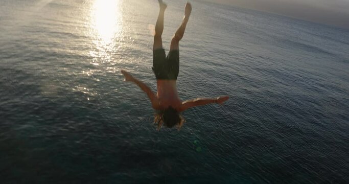 Young man doing extreme backflip cliff jumping backflip the ocean at sunset with lens flare - Powered by Adobe