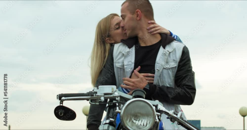 Wall mural A young stylish couple of bikers meet and hug. Man and woman sit on a motorcycle standing in a public city parking lot.