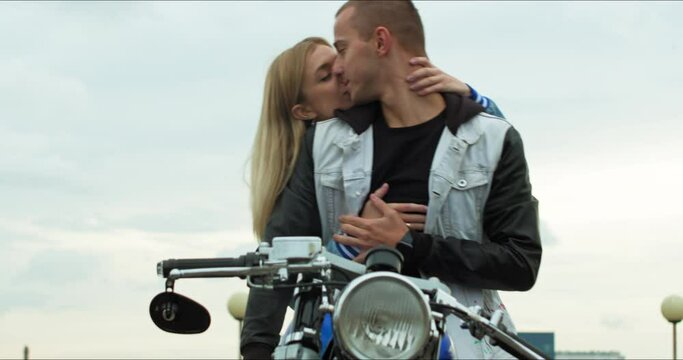 A young stylish couple of bikers meet and hug. Man and woman sit on a motorcycle standing in a public city parking lot.