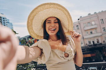 Playful young woman in elegant hat making selfie while relaxing on the balcony