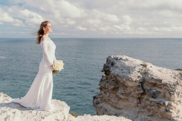 Fototapeta na wymiar Romantic bride, blond girl in white wedding dress with open shoulders posing with sea and rocks in background. Stylish young woman standing on cliff's edge and looking into a ocean mountains at sunset