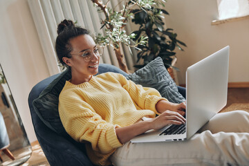Confident young woman using laptop and smiling while sitting in a comfortable chair at home