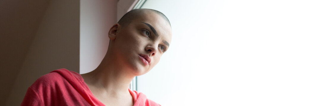 Young depressed female cancer patient standing in hospital beside window
