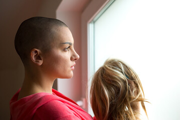 Young depressed female cancer patient standing in hospital beside window