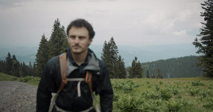 Hiker walking on a macadam road with a backpack, wearing a wind and rain proof jacket. Cloudy weather, wet greens and some spruce trees. Lightlly visible mountains in the backgroud.