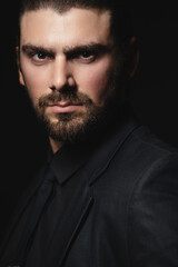 Portrait of a white Caucasian male brunet in a black classic suit, black shirt and black tie over a black background. Fashionable portrait of an attractive man with dark hair