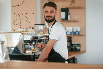 Busy by working with coffee machine. Cafe worker in white shirt and black apron is indoors